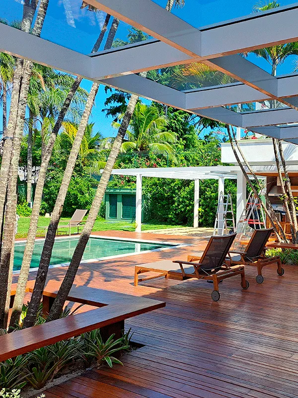 área de lazer com deck de madeira e pergolado branco com cobertura de vidro