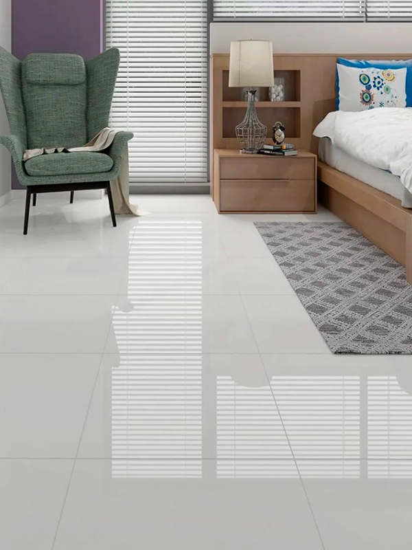 Quarto moderno com cama de madeira, mesa lateral, abajur e poltrona cinza perto de uma janela com persianas projetando sombras em um piso branco brilhante.