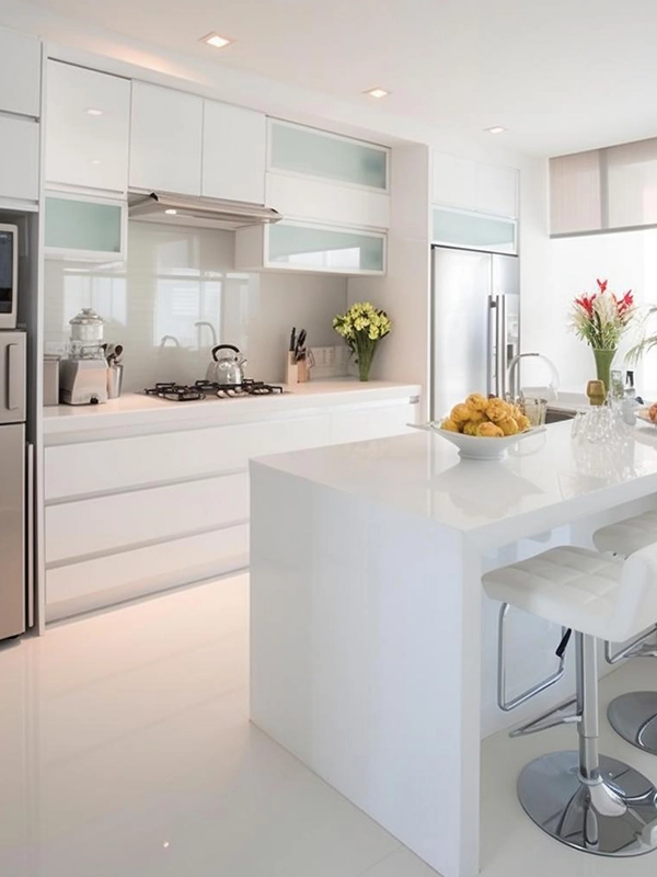 Cozinha moderna branca com armários elegantes, ilha com bancos altos, eletrodomésticos em inox e vaso de flores na bancada com revestimento branco.