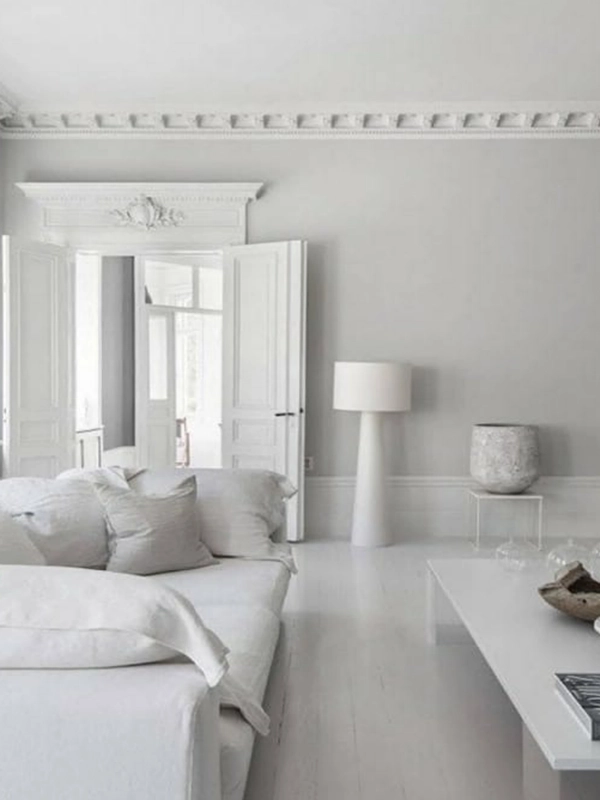 Uma sala minimalista branca com elegante revestimento branco, com sofá branco, mesa de centro, luminária decorativa e portas que dão para outro cômodo.