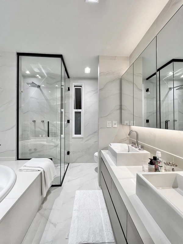 Banheiro moderno com paredes de mármore branco e revestimento branco no piso, com box de vidro, espelho grande e pia branca.