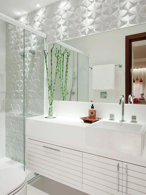 Banheiro branco moderno com parede de azulejos geométricos, porta de box de vidro, pia com penteadeira e vaso com varas de bambu verdes. O espaço é realçado pelo revestimento branco, realçando o seu aspecto clean e