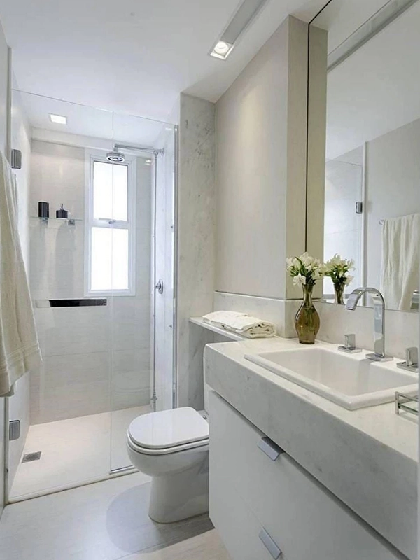 Banheiro moderno com esquema de cores revestimento branco, com pia, espelho, vaso sanitário e box de vidro. As toalhas estão penduradas ordenadamente e um vaso de flores está na bancada.