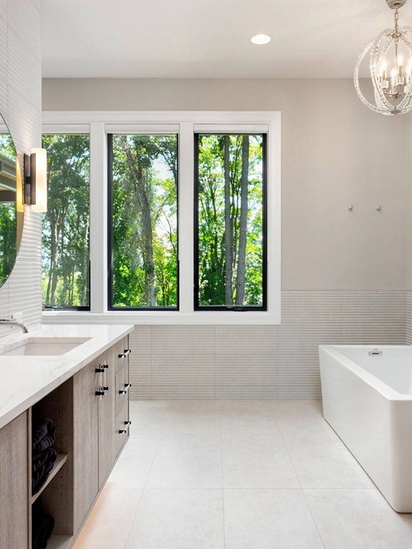 Banheiro moderno com ampla janela com vista para uma exuberante floresta verde, com banheira branca, revestimento branco e armários de madeira.