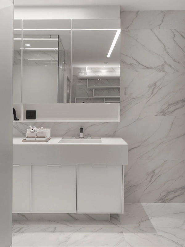 Banheiro moderno com paredes em mármore branco e piso em revestimento branco, pia branca com armários e espelho com iluminação integrada.