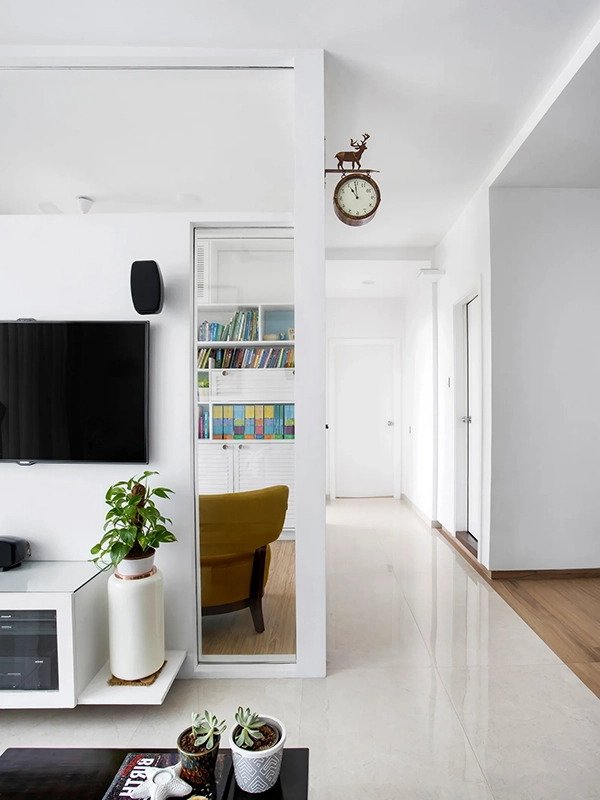 Sala de estar moderna com unidade de entretenimento branca, TV grande e vários vasos de plantas. Uma porta de correr com revestimento branco revela uma estante no cômodo ao lado.