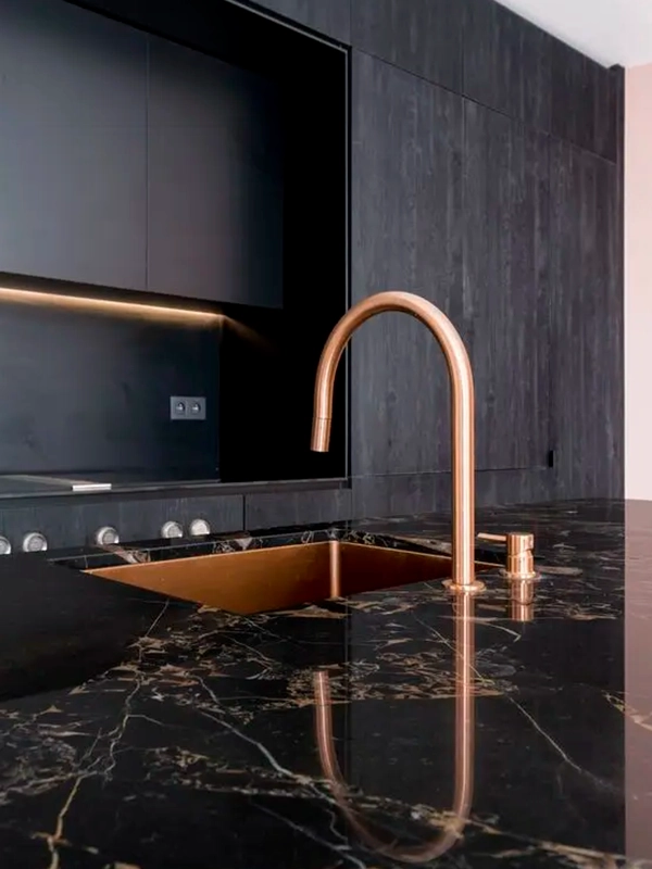 Uma cozinha elegante com torneira e pia em ouro rosa sobre bancada de mármore preto, complementada por armários pretos combinando com revestimento preto e TV embutida.
