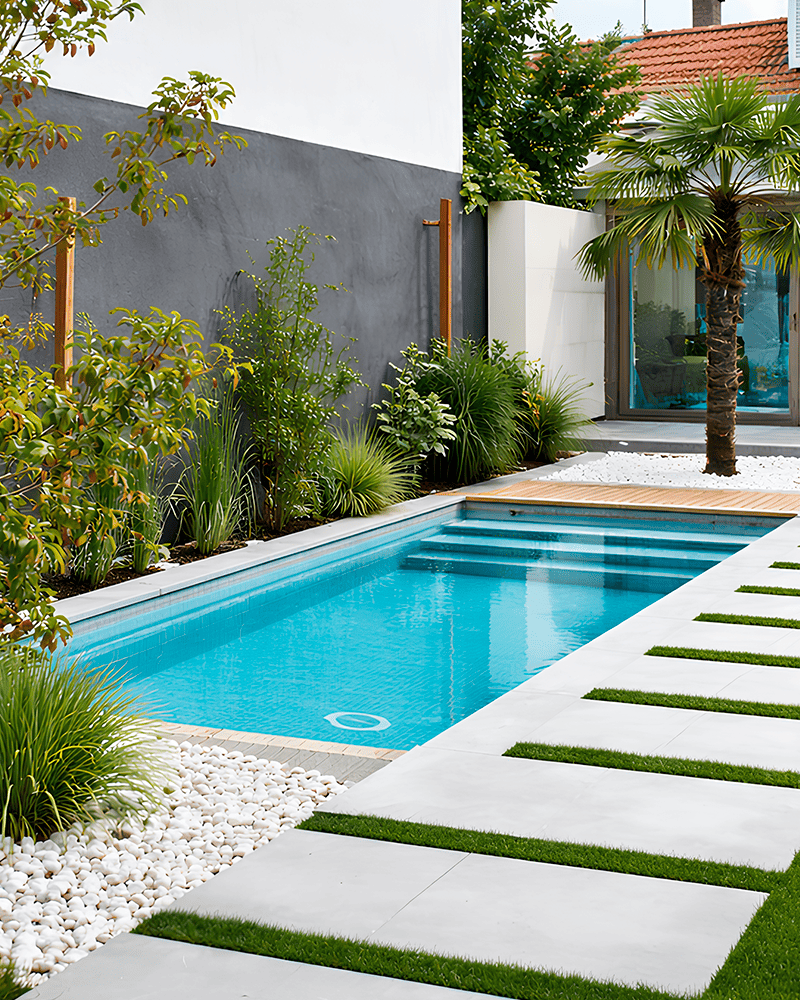 Paisagismo para Piscina: 20 Ideias e Dicas +90 Fotos para Inspirar seu Projeto 219 Piscina retangular no quintal com água limpa, cercada por plantas verdes, seixos e uma palmeira, ao lado de um prédio moderno com grandes portas de vidro.