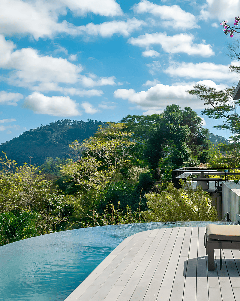 Paisagismo para Piscina: 20 Ideias e Dicas +90 Fotos para Inspirar seu Projeto 220 Piscina infinita e deck de madeira com espreguiçadeira com vista para árvores verdes e uma montanha distante sob um céu azul com nuvens dispersas.