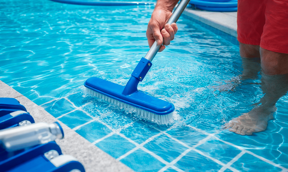 Uma pessoa em pé em uma piscina usa uma escova azul em um poste para esfregar o piso da piscina perto da borda.