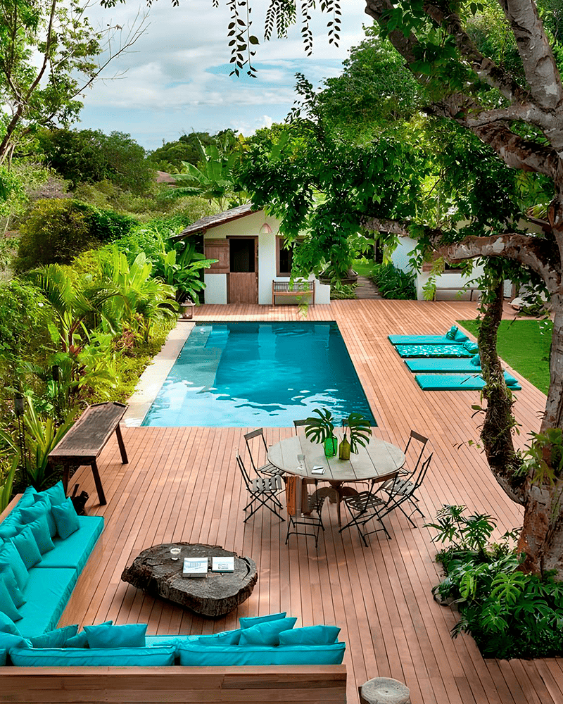 Paisagismo para Piscina: 20 Ideias e Dicas +90 Fotos para Inspirar seu Projeto 221 O quintal apresenta uma piscina retangular, deck de madeira, assentos ao ar livre com almofadas turquesa, uma mesa de jantar e árvores e plantas verdes exuberantes.