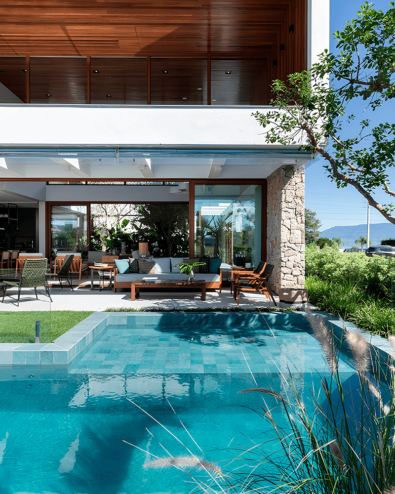 Paisagismo para Piscina: 20 Ideias e Dicas +90 Fotos para Inspirar seu Projeto 223 Casa moderna com teto de madeira, portas de vidro e uma parede de pedra com vista para uma piscina azul clara cercada por vegetação e assentos ao ar livre.