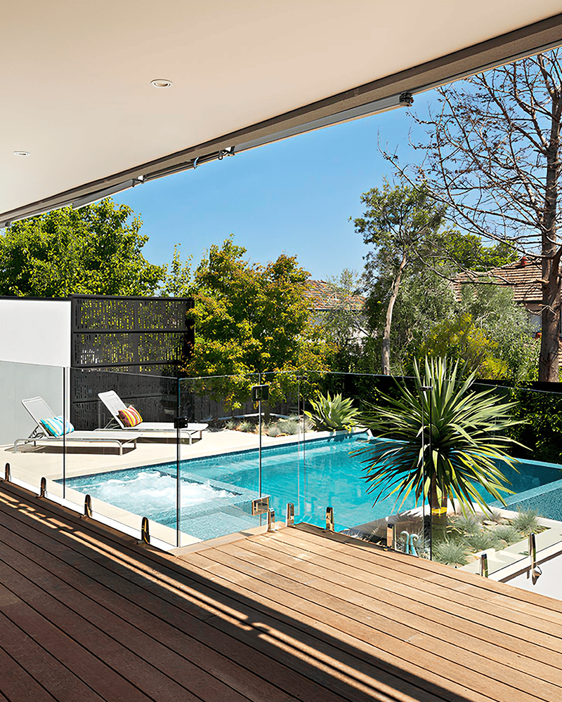 Paisagismo para Piscina: 20 Ideias e Dicas +90 Fotos para Inspirar seu Projeto 224 Um quintal moderno com piscina, espreguiçadeiras, vegetação e um deck de madeira com guarda-corpo de vidro sob um pátio coberto.