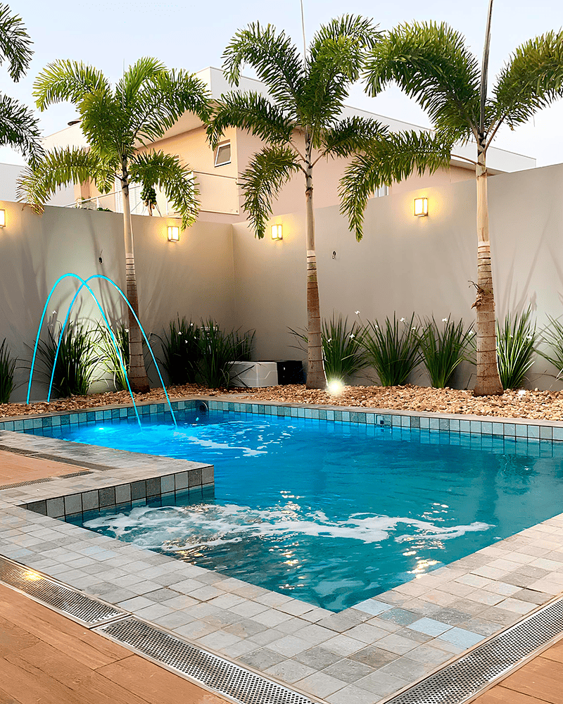 Paisagismo para Piscina: 20 Ideias e Dicas +90 Fotos para Inspirar seu Projeto 225 Uma moderna piscina externa com água azul, uma pequena fonte, deck de azulejos, palmeiras e luzes montadas na parede, cercada por um muro de privacidade.