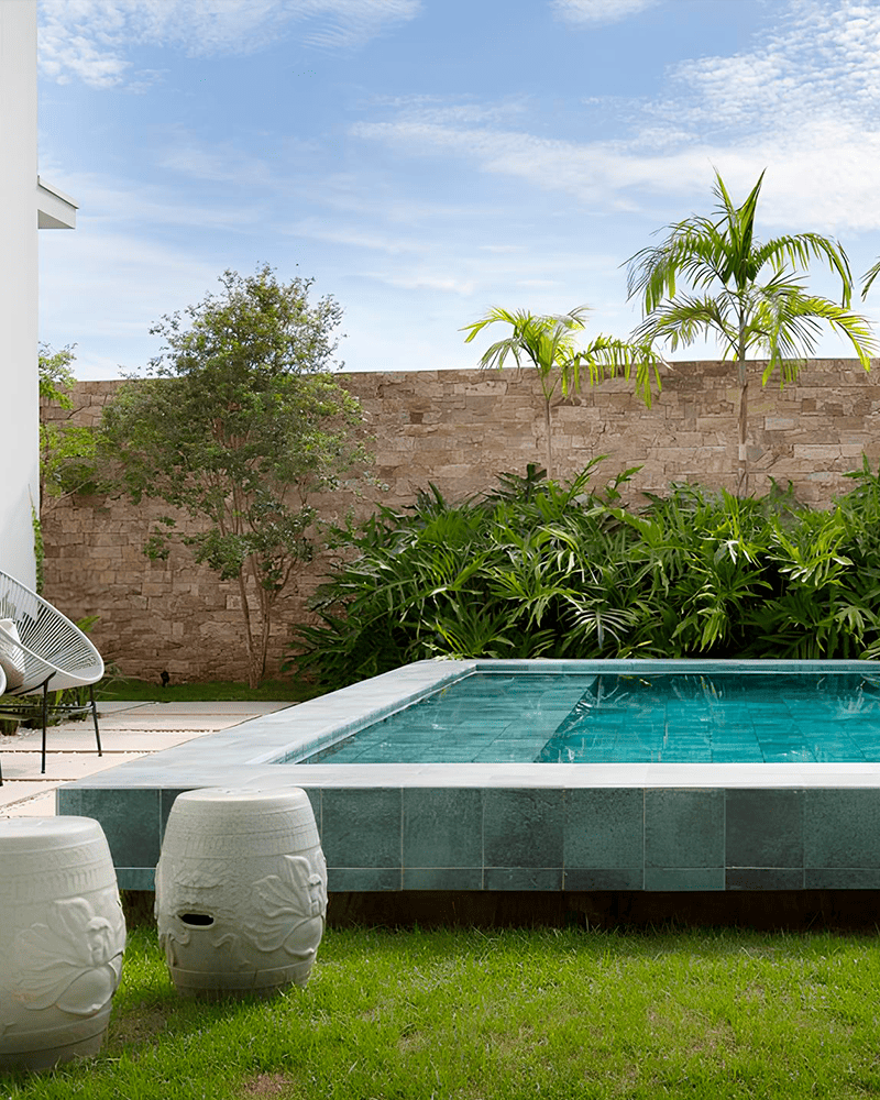 Paisagismo para Piscina: 20 Ideias e Dicas +90 Fotos para Inspirar seu Projeto 226 Um quintal moderno com uma piscina de azulejos, grama verde, móveis de pátio, plantas tropicais e um muro de pedra sob um céu parcialmente nublado.