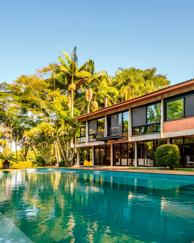 Paisagismo para Piscina: 20 Ideias e Dicas +90 Fotos para Inspirar seu Projeto 227 Casa moderna de dois andares com grandes janelas de vidro cercada por árvores tropicais, com vista para uma piscina azul clara em um dia ensolarado.