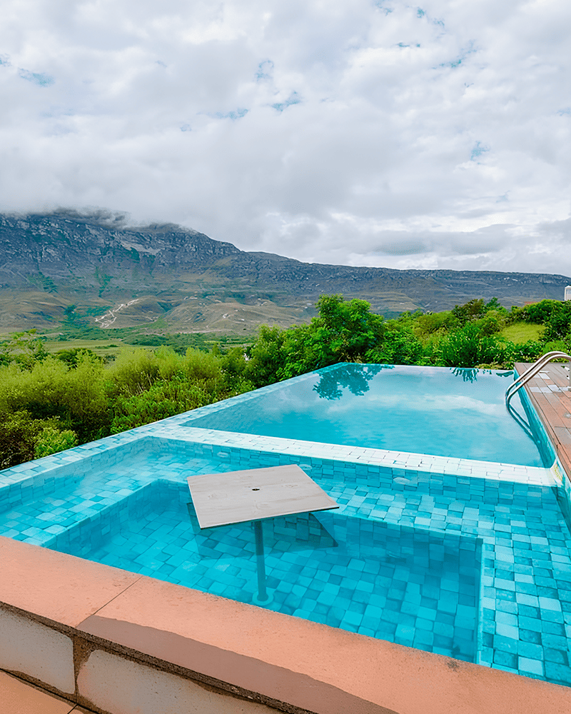 Paisagismo para Piscina: 20 Ideias e Dicas +90 Fotos para Inspirar seu Projeto 230 A piscina infinita com mesa embutida tem vista para as colinas e montanhas verdes sob um céu nublado.