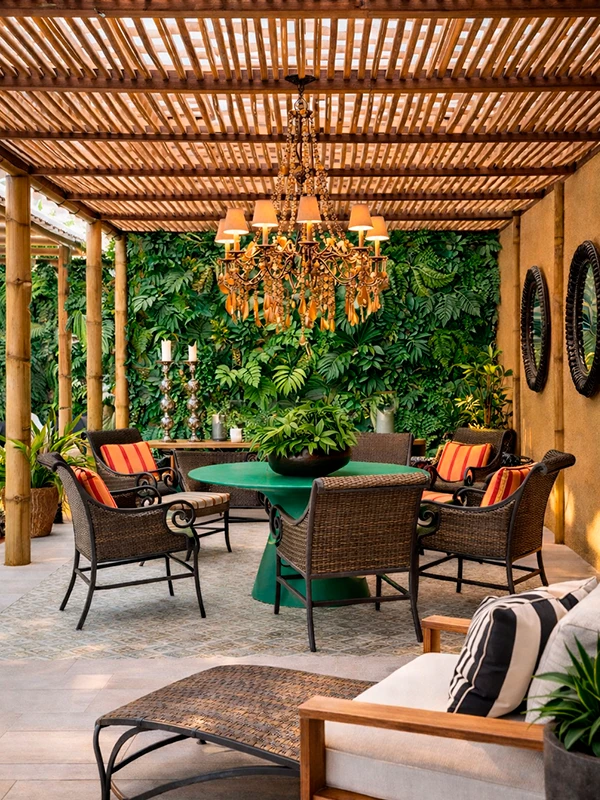 A Área de Lazer com Pergolado conta com uma charmosa estrutura de madeira, cadeiras de vime ao redor de uma mesa verde, almofadas listradas, lustre elegante, jardim vertical exuberante e espelhos decorativos na parede com jardim vertical.