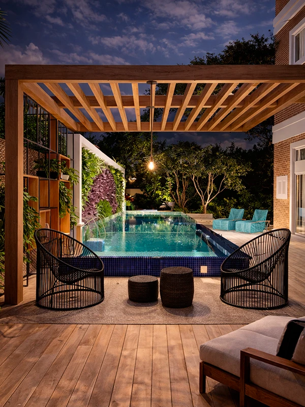 área de estar externa com duas cadeiras pretas sob uma pérgola com vista para uma piscina moderna, cercada por plantas e espreguiçadeiras, lindamente iluminada ao anoitecer.