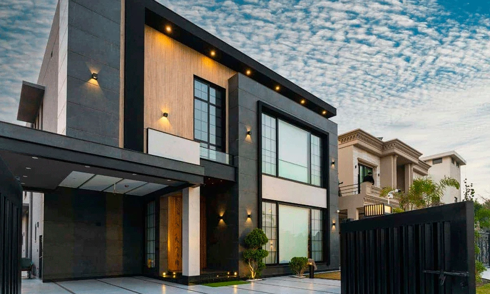 Casa moderna de dois andares com janelas grandes, paredes externas em preto e bege, iluminação externa, jardim paisagístico, portão aberto e porcelanato elegante para garagem sob um céu parcialmente nublado.