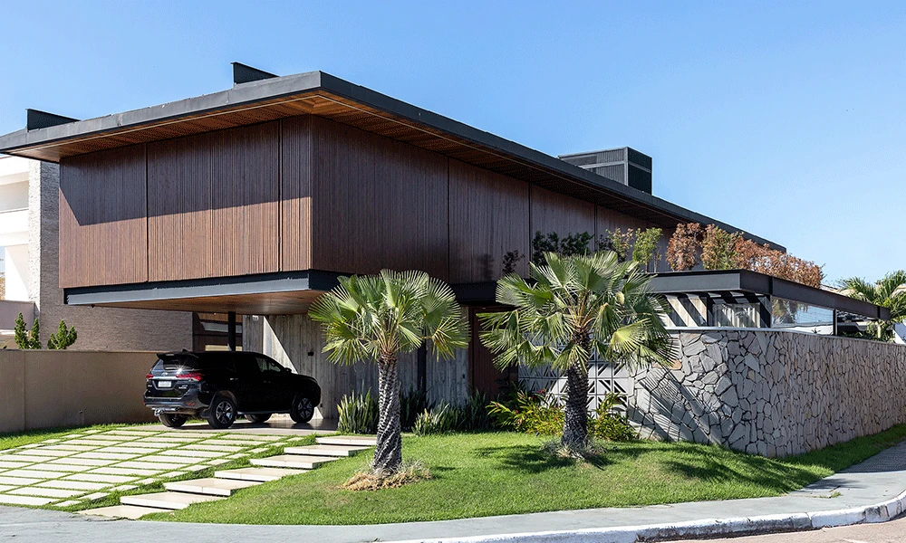 Casa moderna de dois andares com exterior de madeira e pedra, janelas grandes, um SUV preto estacionado na entrada da garagem com porcelanato para garagem e quintal ajardinado com palmeiras.