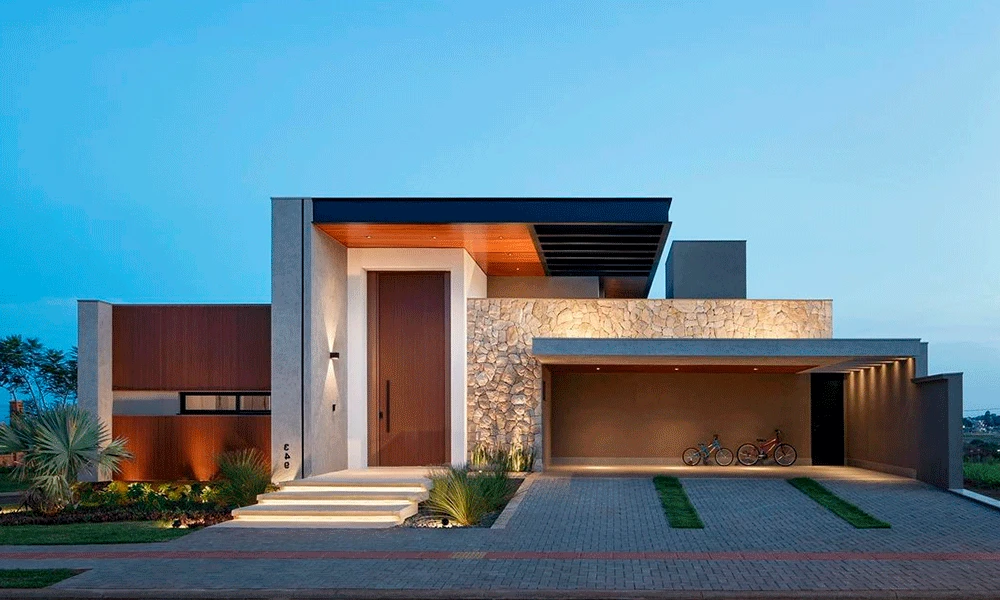 Casa moderna de dois andares com telhado plano, detalhes em pedra e madeira, porta de entrada grande, garagem com duas bicicletas com piso de porcelanato para garagem e um belo jardim paisagístico ao entardecer.