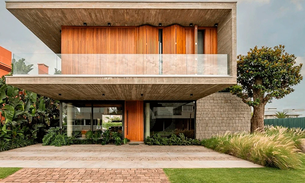 Casa moderna de dois andares com fachada de concreto e madeira, grande varanda de vidro e porcelanato para garagem, cercada por vegetação e um jardim paisagístico.