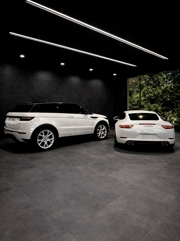 Dois carros brancos, um SUV e um cupê, estão estacionados lado a lado em uma garagem moderna e pouco iluminada, com piso de porcelanato para garagem e uma grande janela com vegetação do lado de fora.
